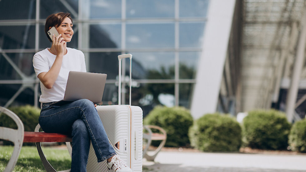 Frau in Jeans und weißem T-Shirt sitzt mit Laptop und Smartphone vor einem Bürogebäude, neben ihr ein Rollkoffer.