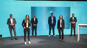 Sechs Personen stehen mit jeweils ca. anderthalb Metern Abstand voneinander in einem Studio vor einem petrolfarbenen Hintergrund mit Grafik