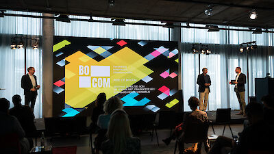 Stefan Rief, Burkhard Kieker und Matthias Schultze stehen auf einer Bühne, vor einem großen Screen mit dem Logo der BOCOM. Von hinten sind im Publikum sitzende Menschen zu sehen.