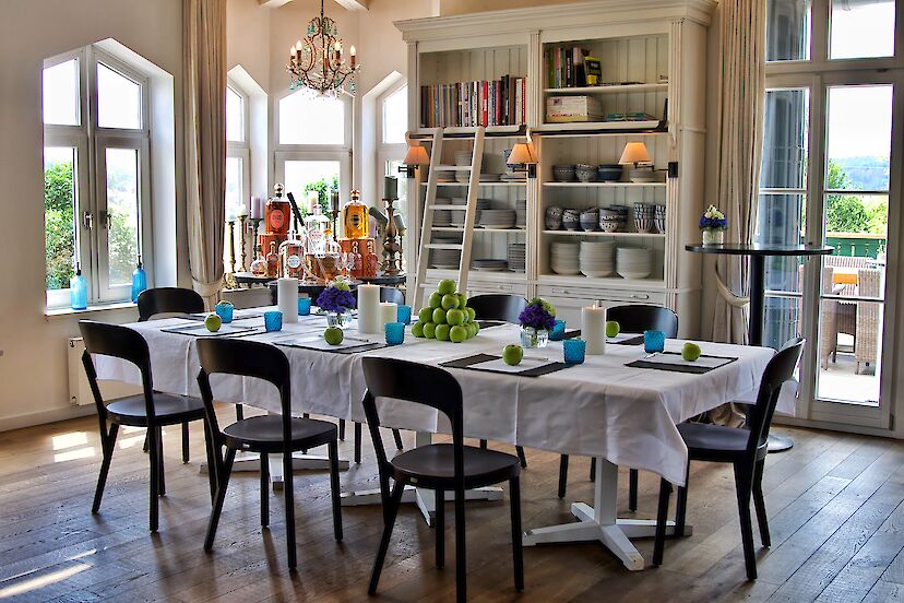 Ein gedeckter Esstisch mit weißem Tischtuch und acht Stühlen in einem Raum im Landhaus-Stil. | © VILA VITA Marburg